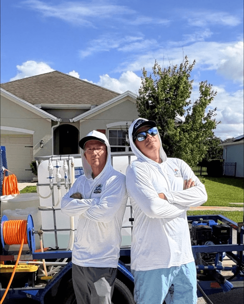 Two Shark Spray technicians standing back to back in front of pressure washing trailer and equipment
